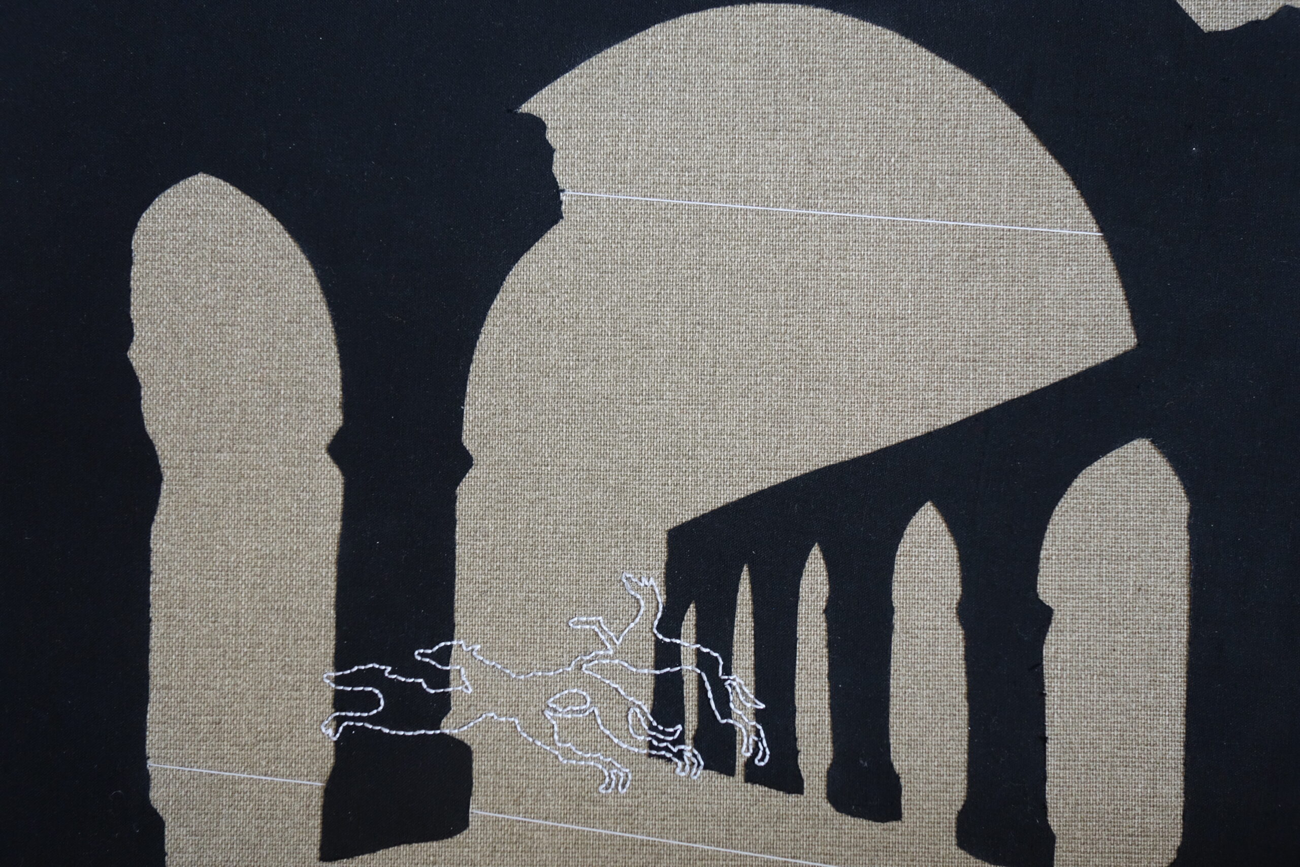 Black silhouettes of arches against a mid brown linen background, with greyhounds outlined in white embroidery