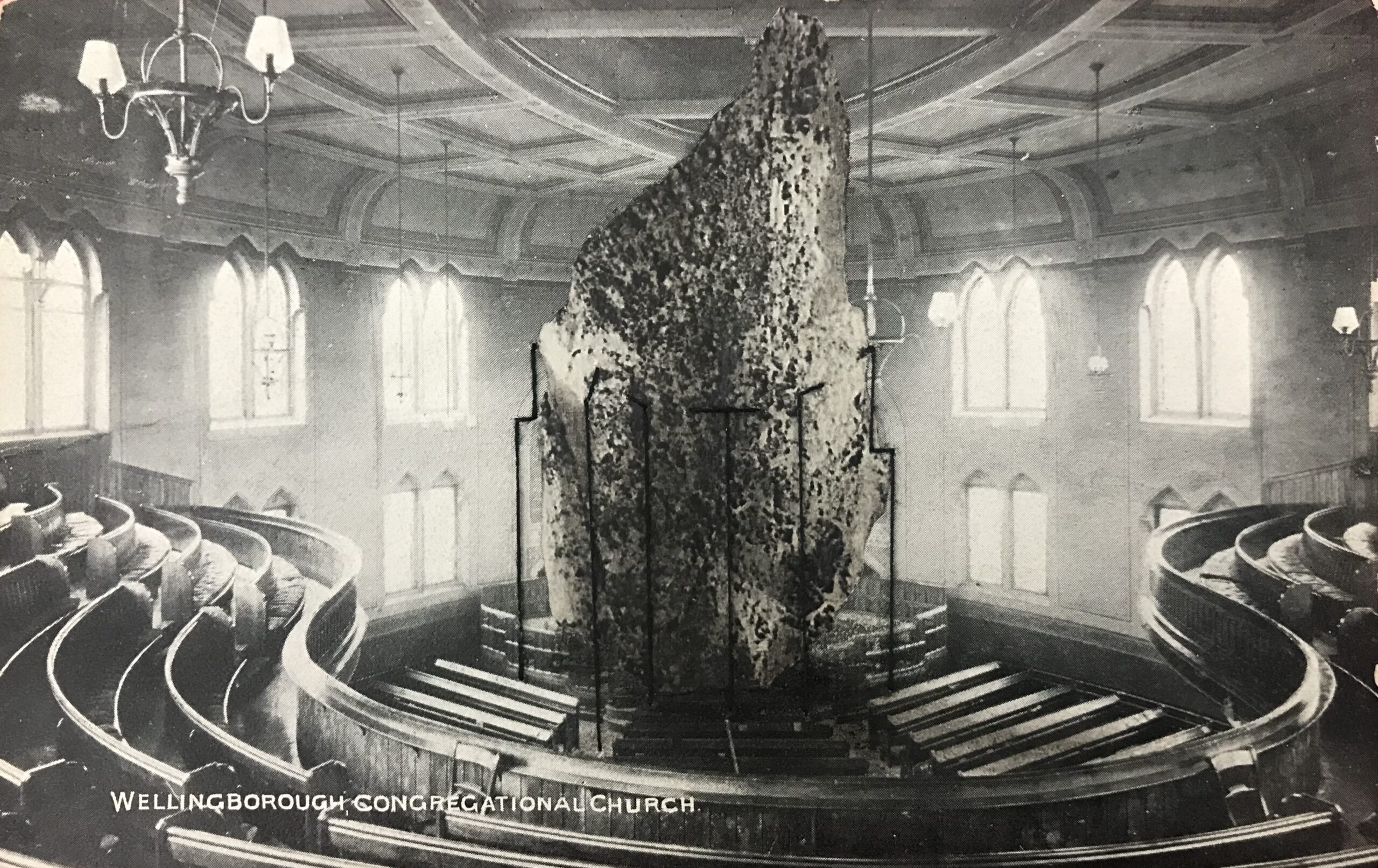Black and white postcard of the inside of a large round building with shaped wooden seating surrounding a central dais. On the dais a huge rock reaches up to the ceiling, held in place with rods made of thread.