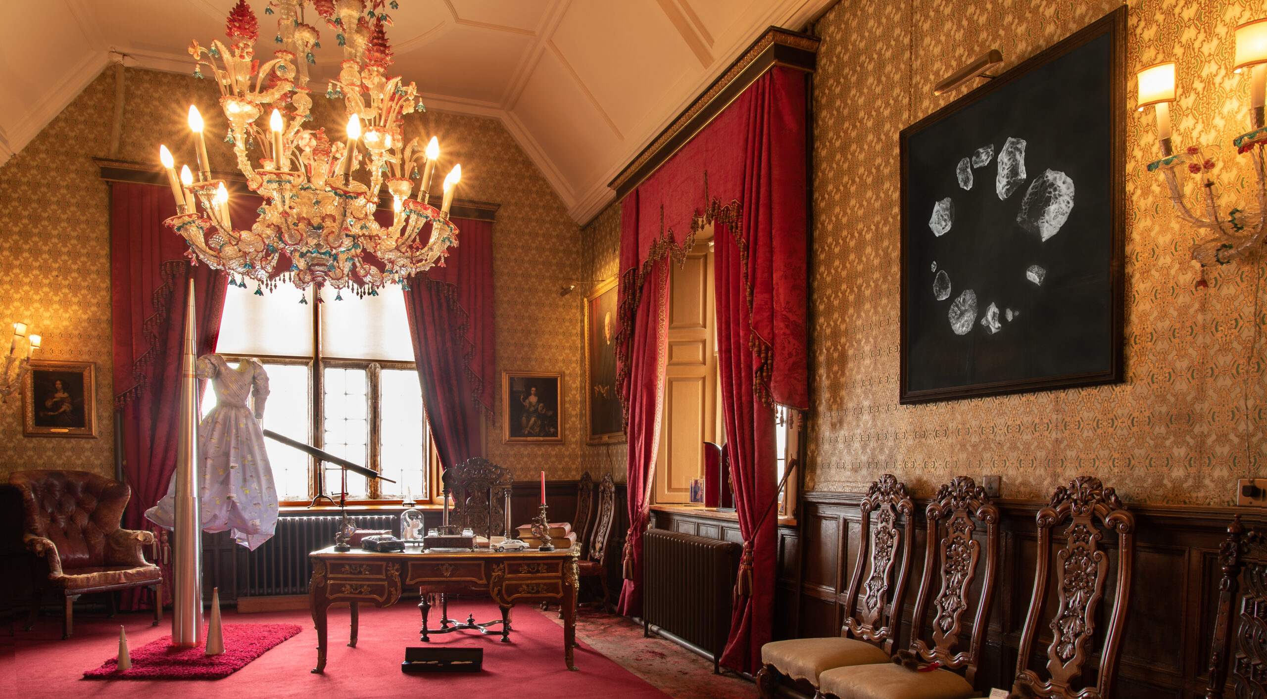 long, grand room inside a stately home, with a red carpet and gold and white wallpaper, fittings and ceiling mouldings. At the far end of the room a dress floats in the air next to a large metal cone; on the right hand wall a drawing on black paper can be seen